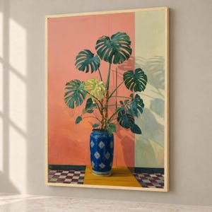 Puede incluir: Un cuadro enmarcado de una gran planta monstera verde en un jarrón de cerámica azul y blanco con motivos. La planta se sitúa sobre una superficie amarilla frente a un fondo dividido en secciones rosa coral y amarillo pálido.