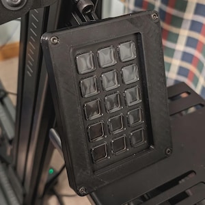 May include: A black 3D-printed macro keypad featuring a rectangular frame with twelve square, translucent keys arranged in a four-by-three grid. The device is mounted on a black metal support structure with visible screws at each corner of the frame.
