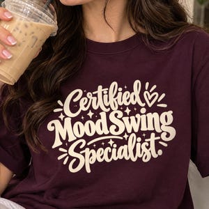 May include: A dark maroon crewneck t-shirt featuring the cream-coloured text "Certified Mood Swing Specialist" printed on the front in a stylised, bold font, accented with small star and heart graphic elements surrounding the lettering.