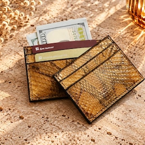 May include: A rectangular card holder featuring a textured, golden-brown snakeskin pattern. The accessory includes multiple slots containing a one-hundred-dollar bill and two credit cards, one of which displays the Cyrillic text Для выплат.