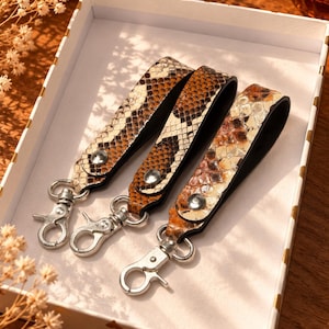 May include: Three wristlet keychains featuring a brown and cream snakeskin pattern. Each strap is secured with a silver-toned metal snap button and includes a matching silver-toned swivel lobster clasp for attaching keys or other small items.