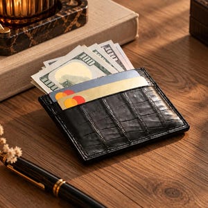 Peut inclure: Un porte-cartes en cuir noir avec un motif texturé en peau de crocodile. Le portefeuille comporte plusieurs fentes contenant plusieurs billets de cent dollars américains et deux cartes de crédit avec des logos circulaires rouges et jaunes.