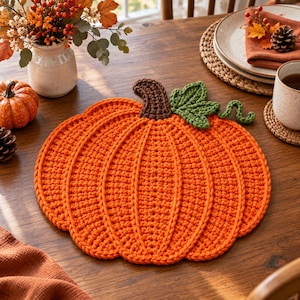 Puede incluir: Un mantel individual redondo de ganchillo naranja con forma de calabaza, que presenta un tallo marrón oscuro y una hoja verde con un zarcillo rizado. El accesorio textil texturizado hecho a mano muestra un patrón acanalado que irradia desde el centro.