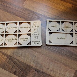 May include: Two wooden boards with hexagonal cutouts, each labeled with different locations and features of a board game. The first board has labels like "Fairy Pool", "Gold Stream", "Hot Springs", "On River", "On Lake", "Wishing Well", "Harvest Spirits Shrine", "Mushroom Grotto", and "Adjacent to Lake". The second board has labels like "Gazebo", "Stone Collector", "Robbers' Lair", "Dormitory", and includes small icons of a cow, a pig, a sheep, a house, and a wheat.