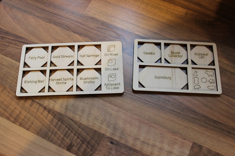 May include: Two wooden boards with hexagonal cutouts, each labeled with different locations and features of a board game. The first board has labels like "Fairy Pool", "Gold Stream", "Hot Springs", "On River", "On Lake", "Wishing Well", "Harvest Spirits Shrine", "Mushroom Grotto", and "Adjacent to Lake". The second board has labels like "Gazebo", "Stone Collector", "Robbers' Lair", "Dormitory", and includes small icons of a cow, a pig, a sheep, a house, and a wheat.