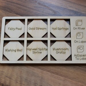 May include: A wooden board with nine hexagonal tiles, each with a different name or symbol. The tiles are labeled "Fairy Pool", "Gold Stream", "Hot Springs", "On River", "On Lake", "Wishing Well", "Harvest Spirits Shrine", "Mushroom Grotto", and "Adjacent To Lake".