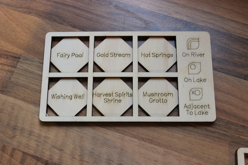 May include: A wooden board with nine hexagonal tiles, each with a different name or symbol. The tiles are labeled "Fairy Pool", "Gold Stream", "Hot Springs", "On River", "On Lake", "Wishing Well", "Harvest Spirits Shrine", "Mushroom Grotto", and "Adjacent To Lake".