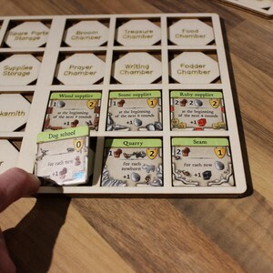 May include: A wooden game board with hexagonal spaces for holding game pieces. The board is filled with various game pieces, including a dog school card, a quarry card, and a seam card. The cards have numbers and text on them, indicating their value and function in the game.