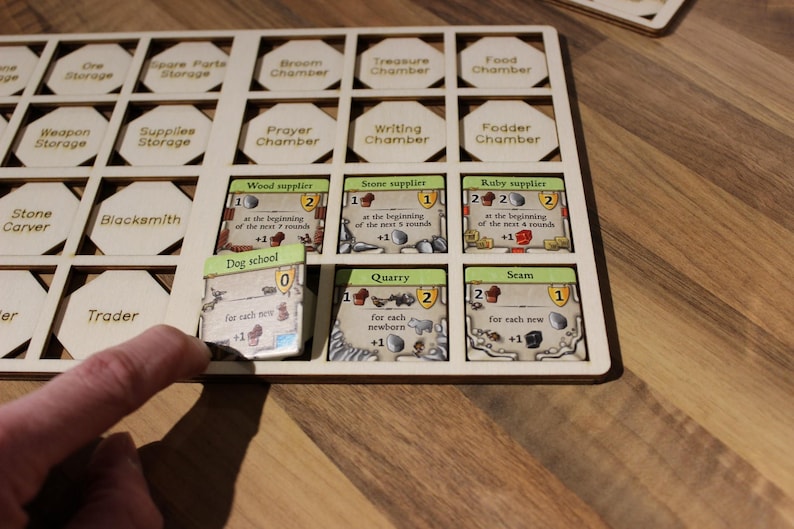 May include: A wooden game board with hexagonal spaces for holding game pieces. The board is filled with various game pieces, including a dog school card, a quarry card, and a seam card. The cards have numbers and text on them, indicating their value and function in the game.