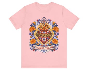 Może przedstawiać: Jasnoróżowy t-shirt z centralną grafiką Najświętszego Serca otoczonego pomarańczowymi kwiatami nagietka, niebieskimi liśćmi i krzyżem. Serce jest otoczone koroną cierniową, a poniżej znajduje się wstęga z napisem SAGRADO CORAZÓN.