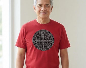 Può includere: Una t-shirt rossa a maniche corte con una grafica circolare della medaglia di San Benedetto al centro. Il design include una croce con le lettere C S S M L e N D S M D, circondata dall'iscrizione PAX e da varie abbreviazioni latine.
