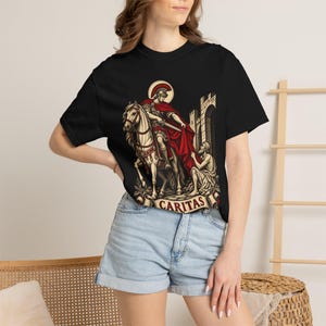 Puede incluir: Una camiseta negra de manga corta con un estampado gráfico de un soldado romano a caballo con capa roja y espada, compartiendo su manto con una figura arrodillada junto a un muro de piedra. El diseño incluye el texto CARITAS.