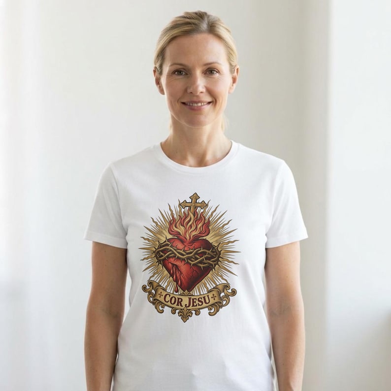 K&ouml;nnte beinhalten: Ein wei&szlig;es T-Shirt mit einer Grafik eines roten Herzens, das von einer Dornenkrone umgeben ist, gekr&ouml;nt von einem Kreuz und strahlenden Lichtstrahlen. Unter dem Herzen befindet sich ein dekoratives Banner mit dem Text COR JESU.