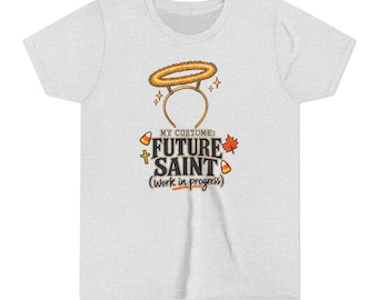 May include: A light grey short-sleeve t-shirt featuring a graphic of a halo headband above the text "MY COSTUME: FUTURE SAINT (work in progress)". The design includes small candy corn, a cross, and an orange maple leaf illustration.