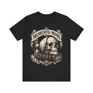 Puede incluir: Una camiseta negra con un estampado gr&aacute;fico de un cr&aacute;neo humano sobre un libro, acompa&ntilde;ado de un reloj de arena y un rosario de cuentas rojas. Una cinta sobre el cr&aacute;neo muestra el texto MEMENTO MORI en una fuente decorativa.