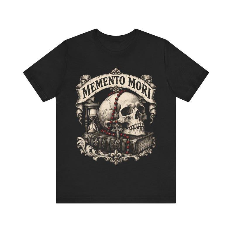 Puede incluir: Una camiseta negra con un estampado gr&aacute;fico de un cr&aacute;neo humano sobre un libro, acompa&ntilde;ado de un reloj de arena y un rosario de cuentas rojas. Una cinta sobre el cr&aacute;neo muestra el texto MEMENTO MORI en una fuente decorativa.