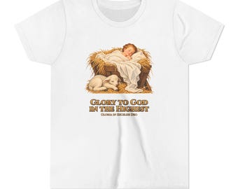 May include: A white short-sleeve t-shirt featuring a graphic of a baby sleeping in a manger with a lamb. Below the illustration, the text reads "GLORY TO GOD IN THE HIGHEST" and "GLORIA IN EXCELSIS DEO" in brown, serif capital letters.