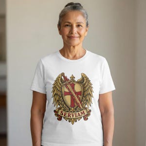 Op de afbeelding: Een wit T-shirt met korte mouwen en een centrale afbeelding van een gouden schild met een rood kruis, een vlammend zwaard en grote vleugels. Onder het schild staat een rode banner met het woord SERVIAM in gouden letters.