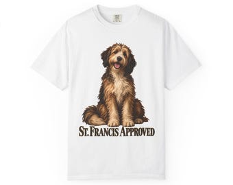 Può includere: Una t-shirt bianca a maniche corte con la grafica di un cane a pelo lungo marrone e nero seduto. Sotto il cane, il testo "ST. FRANCIS APPROVED" è stampato in un carattere serif grassetto marrone scuro centrato sul petto.