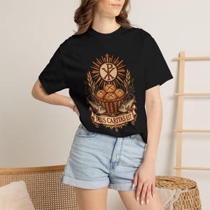 May include: A black short-sleeve t-shirt featuring a graphic print of a basket of bread and two fish, topped with a Chi-Rho symbol and wheat stalks. Below the basket, a banner displays the Latin text DEUS CARITAS EST in gold lettering.