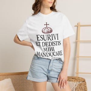 Może przedstawiać: Biały t-shirt z krótkim rękawem z ciemnobrązową grafiką przedstawiającą bochenek chleba z krzyżem. Pod grafiką znajduje się napis ESURIVI ET DEDISTIS MIHI MANDUCARE oraz MT 25:35 wykonany czarną czcionką szeryfową.