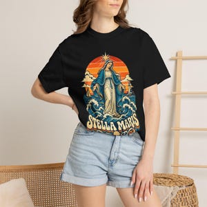 May include: A black short-sleeve t-shirt featuring a graphic print of a figure in a blue and white robe standing on ocean waves. The design includes an orange sun, a star, a sailboat, and the text Stella Maris in a retro font.
