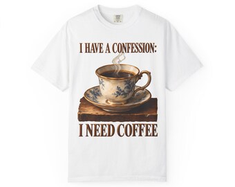 Può includere: Una t-shirt bianca a maniche corte con la grafica di una tazza di caffè fumante su un piattino appoggiato su un blocco di legno. Il testo sopra recita I HAVE A CONFESSION: e il testo sotto I NEED COFFEE in lettere marroni.