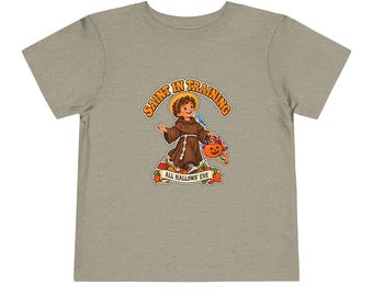 May include: A heathered light brown t-shirt featuring a graphic of a child dressed as a monk holding a jack-o'-lantern bucket. The text reads SAINT IN TRAINING above and ALL HALLOWS' EVE below, surrounded by colorful autumn leaves.