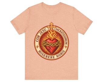 Puede incluir: Una camiseta de manga corta color melocotón con un gráfico circular de un Sagrado Corazón rojo rodeado por una corona de espinas, coronado con una cruz y llamas, con el texto COR JESU SACRATISSIMUM MISERERE NOBIS.