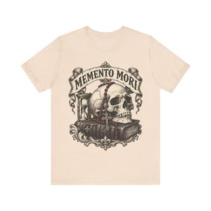Puede incluir: Una camiseta de color beige claro con un estampado gr&aacute;fico de un cr&aacute;neo humano sobre un libro, acompa&ntilde;ado de un reloj de arena y un rosario de cuentas rojas. Una cinta sobre el cr&aacute;neo muestra el texto MEMENTO MORI en letra oscura.