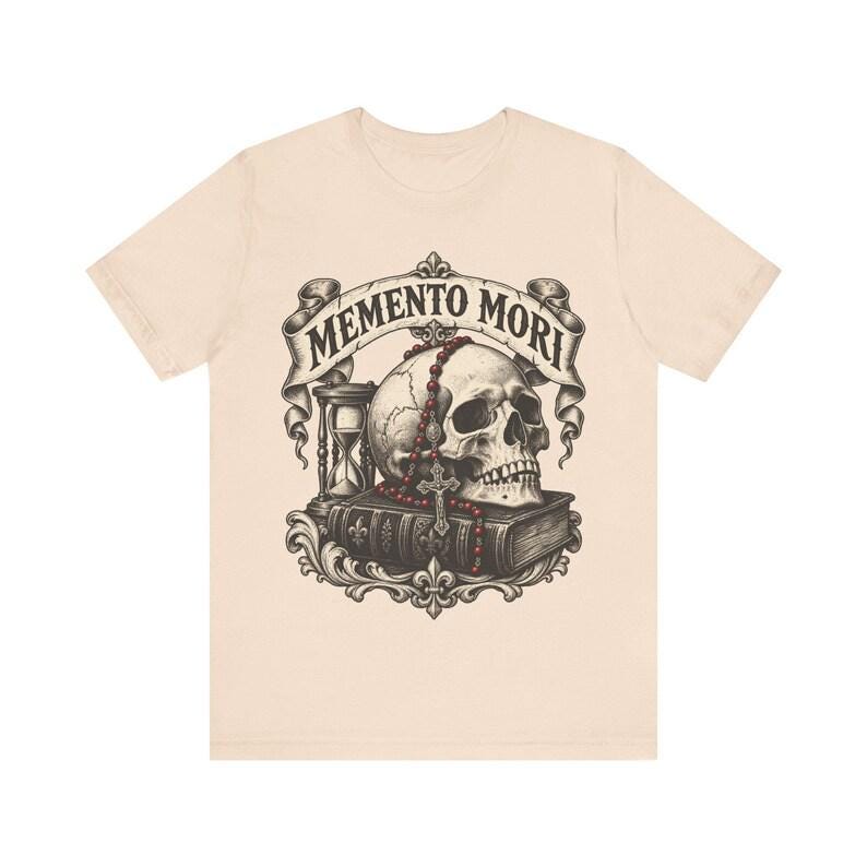 Puede incluir: Una camiseta de color beige claro con un estampado gr&aacute;fico de un cr&aacute;neo humano sobre un libro, acompa&ntilde;ado de un reloj de arena y un rosario de cuentas rojas. Una cinta sobre el cr&aacute;neo muestra el texto MEMENTO MORI en letra oscura.