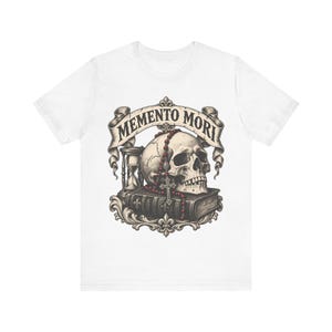 Puede incluir: Una camiseta blanca con un estampado gr&aacute;fico de un cr&aacute;neo humano sobre un libro, acompa&ntilde;ado de un reloj de arena y un rosario de cuentas rojas. Sobre el cr&aacute;neo, una cinta muestra el texto MEMENTO MORI en una fuente oscura y definida.