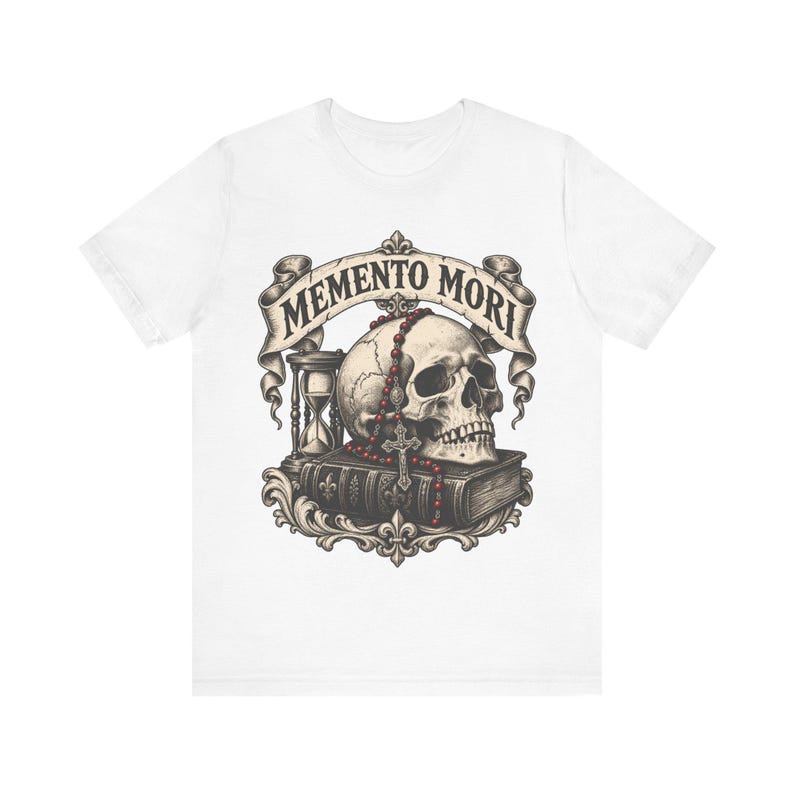 Puede incluir: Una camiseta blanca con un estampado gr&aacute;fico de un cr&aacute;neo humano sobre un libro, acompa&ntilde;ado de un reloj de arena y un rosario de cuentas rojas. Sobre el cr&aacute;neo, una cinta muestra el texto MEMENTO MORI en una fuente oscura y definida.