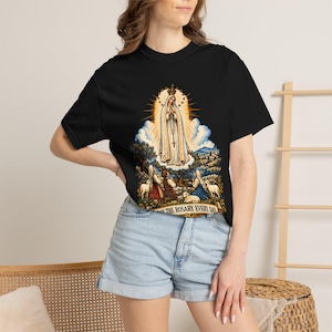 以下が含まれることがあります： 黒の半袖Tシャツ。白いローブをまとった人物が金色の光輪、雲、羊を連れた跪く人々に囲まれているグラフィックプリントが施されている。イラストの下のバナーには「PRAY THE ROSARY EVERY DAY」というテキストが表示されている。