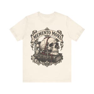 Puede incluir: Una camiseta de color crema con un estampado gr&aacute;fico de un cr&aacute;neo humano sobre un libro, acompa&ntilde;ado de un reloj de arena y un rosario de cuentas rojas. Sobre el cr&aacute;neo, una cinta muestra el texto MEMENTO MORI en letras negras.