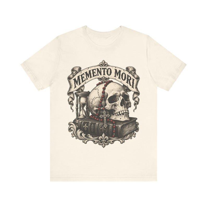 Puede incluir: Una camiseta de color crema con un estampado gr&aacute;fico de un cr&aacute;neo humano sobre un libro, acompa&ntilde;ado de un reloj de arena y un rosario de cuentas rojas. Sobre el cr&aacute;neo, una cinta muestra el texto MEMENTO MORI en letras negras.