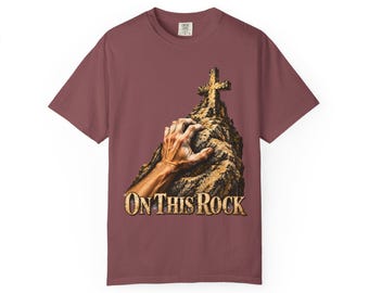 Può includere: Una t-shirt a maniche corte color bordeaux con una grafica che raffigura una mano che raggiunge una croce in cima a una montagna rocciosa. Il testo "ON THIS ROCK" è stampato in un carattere dorato e testurizzato sotto l'immagine.