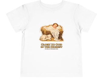 May include: A white t-shirt featuring a graphic of a baby sleeping in a manger filled with hay, with a small lamb resting nearby. Below the illustration, the text reads GLORY TO GOD IN THE HIGHEST and GLORIA IN EXCELSIS DEO in gold lettering.