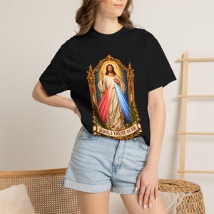 May include: A black short-sleeve t-shirt featuring a central graphic of Jesus Christ within an ornate gold frame. The figure wears white robes with red and blue rays emanating from the chest. Below the image, a banner reads: JESUS, I TRUST IN YOU.