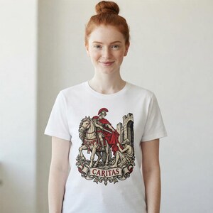 Puede incluir: Una camiseta blanca de manga corta con una ilustraci&oacute;n gr&aacute;fica de un soldado romano a caballo compartiendo una capa roja con una persona sentada junto a un muro de piedra. Debajo de la escena hay una pancarta con la palabra CARITAS.