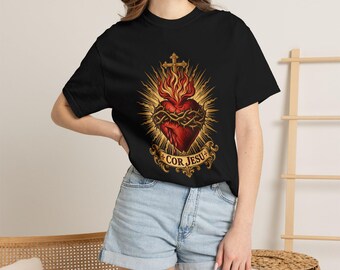 May include: A black short-sleeve t-shirt featuring a graphic of a red Sacred Heart encircled by a crown of thorns, topped with a cross and flames, with a banner below displaying the Latin text COR JESU in gold lettering.