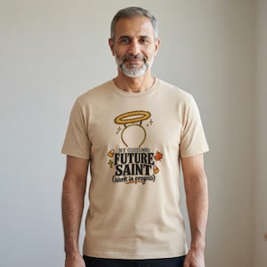 Pu&ograve; includere: Una t-shirt beige a maniche corte con una grafica di un cerchietto con aureola sopra il testo "MY COSTUME: FUTURE SAINT (Work in progress)". Il design include piccole icone di caramelle a forma di mais, una croce e una foglia autunnale arancione.