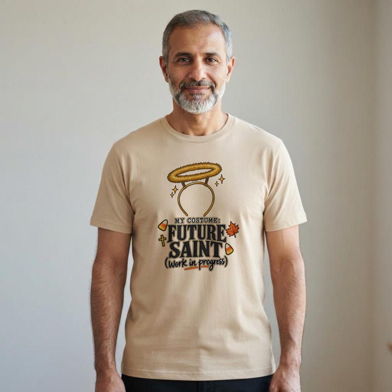 Pu&ograve; includere: Una t-shirt beige a maniche corte con una grafica di un cerchietto con aureola sopra il testo "MY COSTUME: FUTURE SAINT (Work in progress)". Il design include piccole icone di caramelle a forma di mais, una croce e una foglia autunnale arancione.