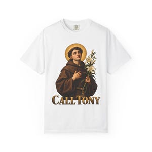 Puede incluir: Una camiseta blanca de manga corta con un estampado gráfico de una figura con túnica marrón con capucha y halo dorado, sosteniendo lirios blancos. Debajo de la figura, el texto CALL TONY está impreso en una fuente serif dorada y negrita.