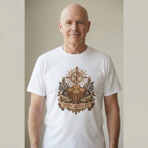 May include: A white short-sleeved t-shirt featuring a central graphic of a basket of bread and two fish beneath a Chi-Rho symbol, wheat stalks, and a banner with the Latin text DEUS CARITAS EST in brown, gold, and tan tones.