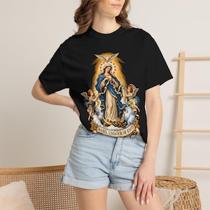 May include: A black short-sleeved t-shirt featuring a graphic print of Mary, Undoer of Knots. The design depicts a figure in blue and white robes surrounded by angels, clouds, and a dove, with the text MARY, UNDOER OF KNOTS at the base.