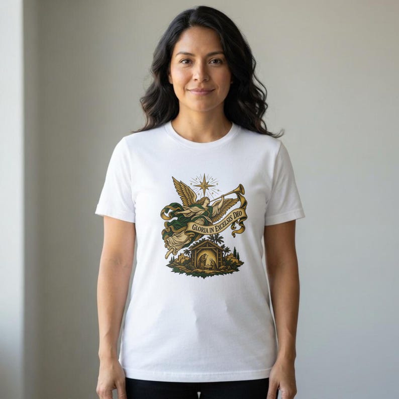 May include: A white short-sleeved t-shirt featuring a central graphic of an angel blowing a trumpet above a nativity scene. A banner draped across the illustration contains the text Gloria in Excelsis Deo in a dark, ornate font.