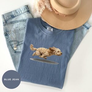 May include: A blue t-shirt featuring a graphic of a running tan dog, paired with light blue denim jeans, a tan wide-brimmed hat, and a gold chain necklace. The shirt has a Comfort Colors label at the inner neckline and a blue jean colour swatch.