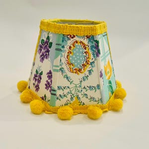 May include: A small lampshade with a floral pattern in shades of blue, purple, and yellow. The lampshade is trimmed with yellow pom-poms and a yellow crocheted edge. The lampshade is approximately 6 inches tall.