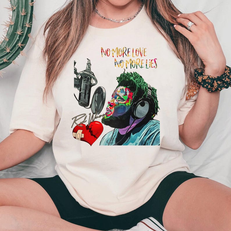 May include: Cream-colored t-shirt featuring a colorful graphic of a musician with headphones and a microphone. The graphic includes the text "NO MORE LOVE NO MORE LIES" and a broken heart. The artist's name is also visible.
